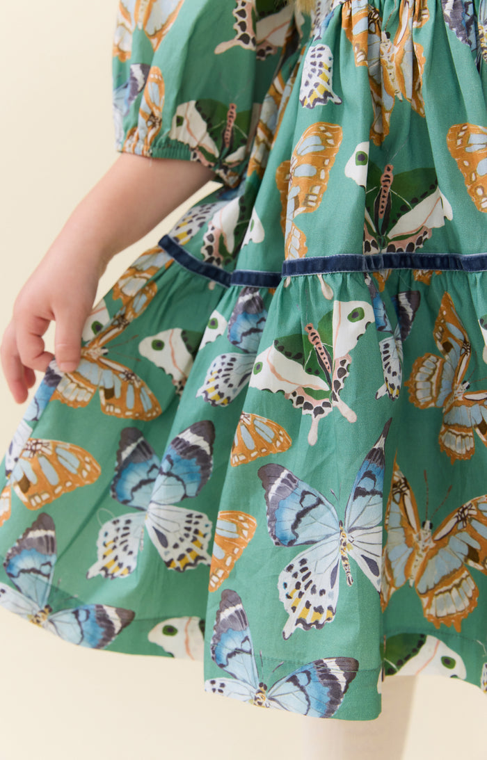 Pink Chicken’s Girls Maribelle Dress in Green Butterflies brings playful elegance to every twirl. The delicate butterfly print and navy velour ribbon trim create a charming look that delights and inspires confidence in every step. Perfect for joyful moments and unforgettable memories!