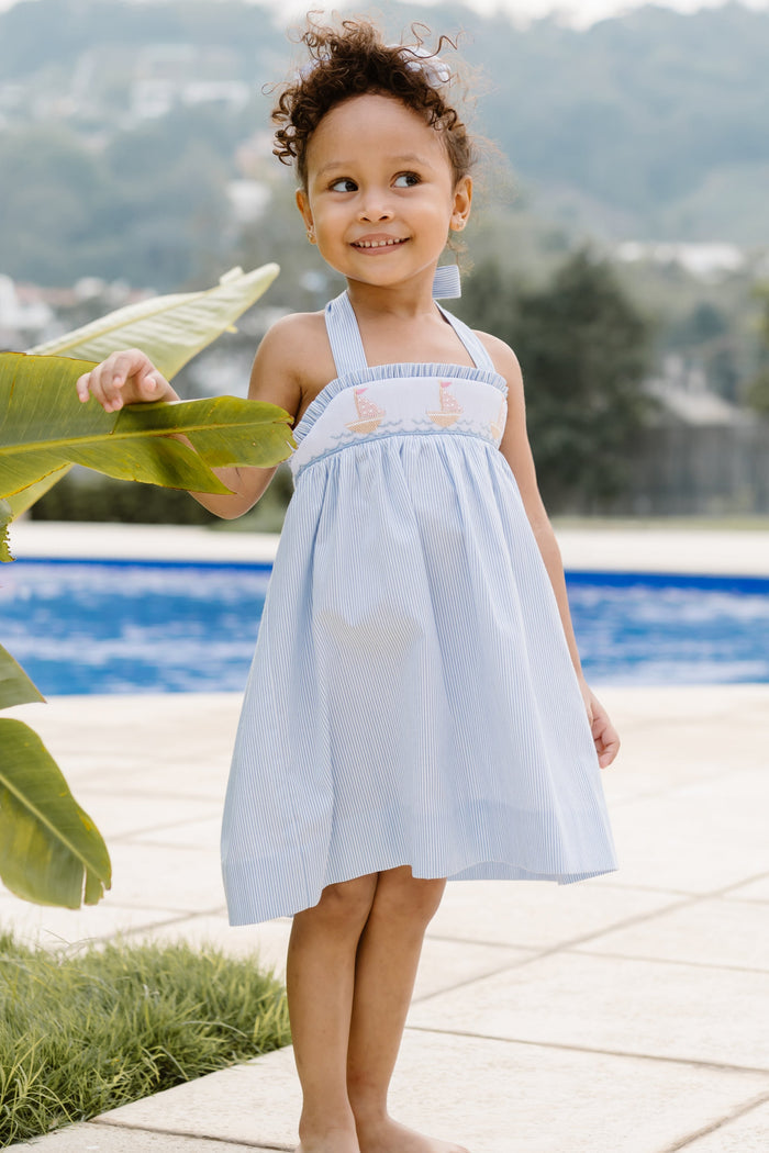 This enchanting Girl's Rachel Sailboat Stripe Dress captures summer's essence with classic blue and white seersucker stripes. The beautifully smocked bodice showcases charming embroidered sailboats, while the halter tie design ensures a perfect fit. An irresistible choice for warm-weather adventures, this quintessential summer dress is available for both toddlers and girls. Complete the look with the matching little sister short set for adorable coordinated outfits!